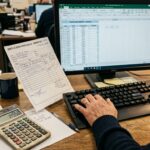 Wholesale office worker entering order data from paper forms into spreadsheet with calculator and invoices on desk